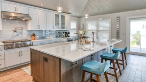 A modern kitchen with white cabinetry, a central island with bar stools, and a large window with blinds. - Brand New! Steps to Beach, Private Pool and Hot Tub, Putt-Putt, Fenced Yard, $300 in Free Tickets! (St. Augustine)