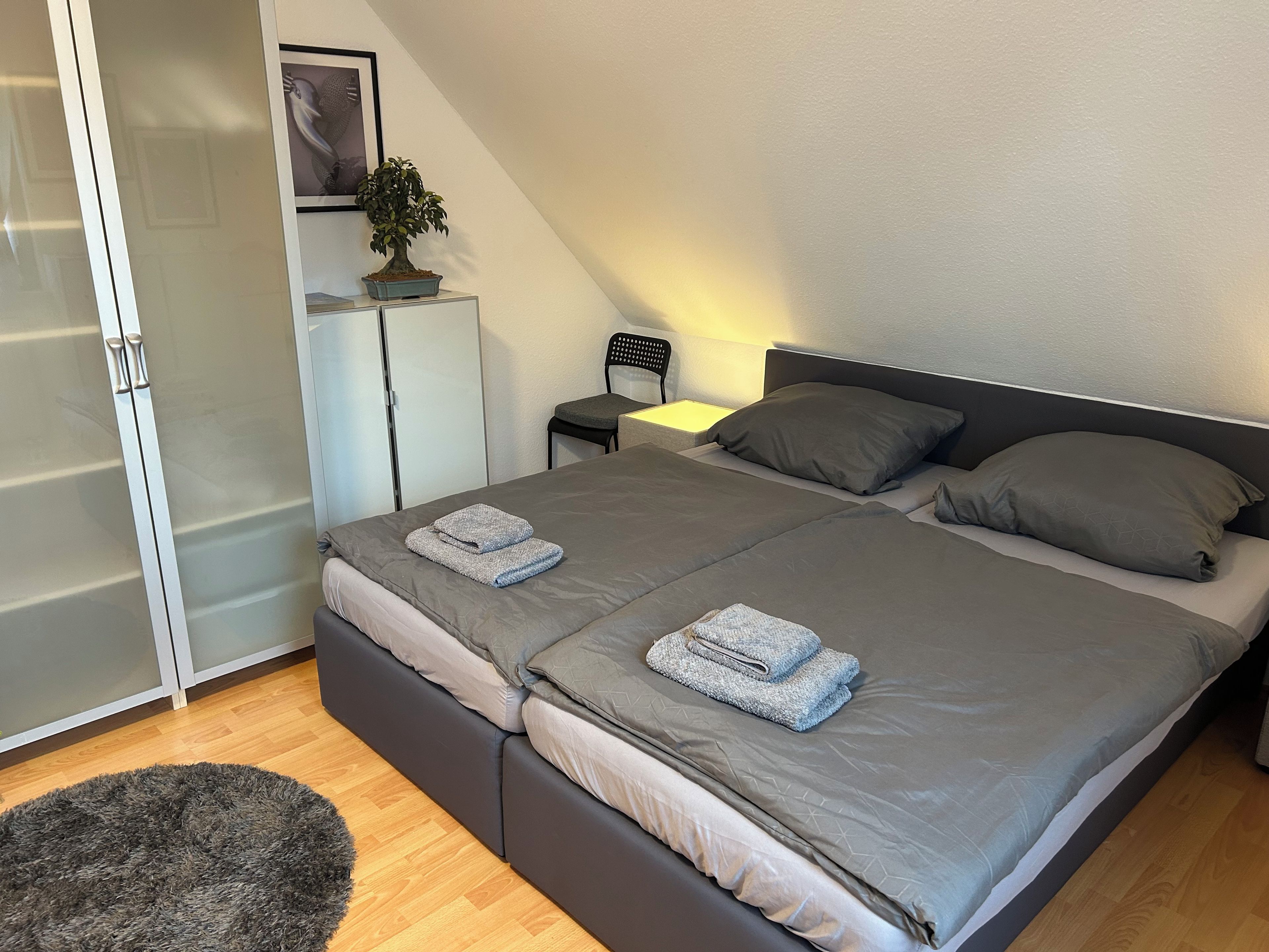 Une chambre à coucher avec un lit, des oreillers, une table de chevet, une chaise, une armoire et une plante en pot.