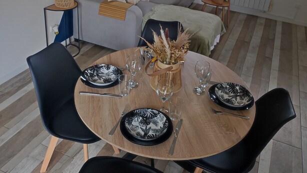 Un coin repas avec une table ronde en bois, des chaises noires et un canapé gris en arrière-plan.