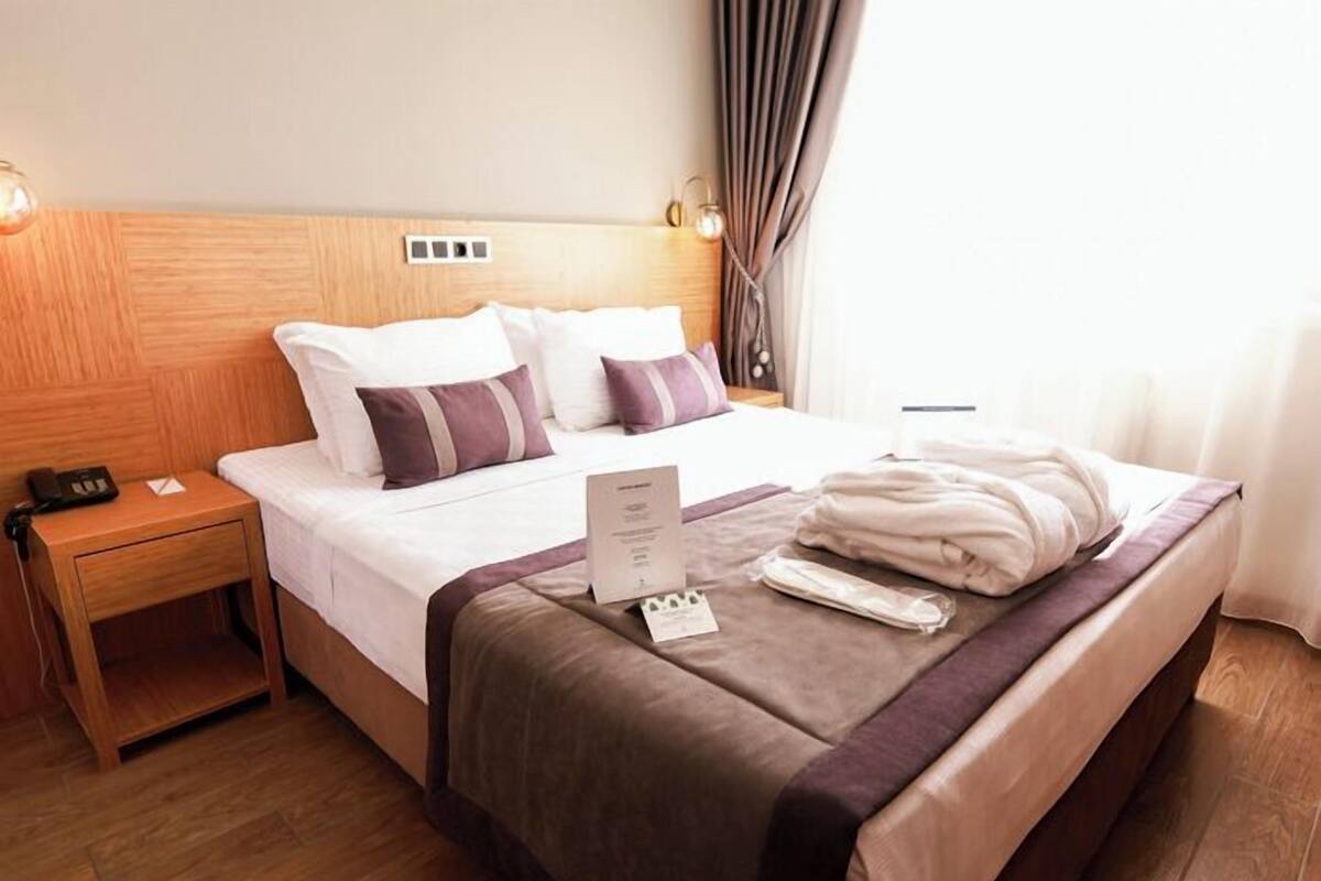 A hotel room with a wooden headboard, a neatly made bed with white and purple bedding, a wooden nightstand with a phone, and a bedside table with a menu.