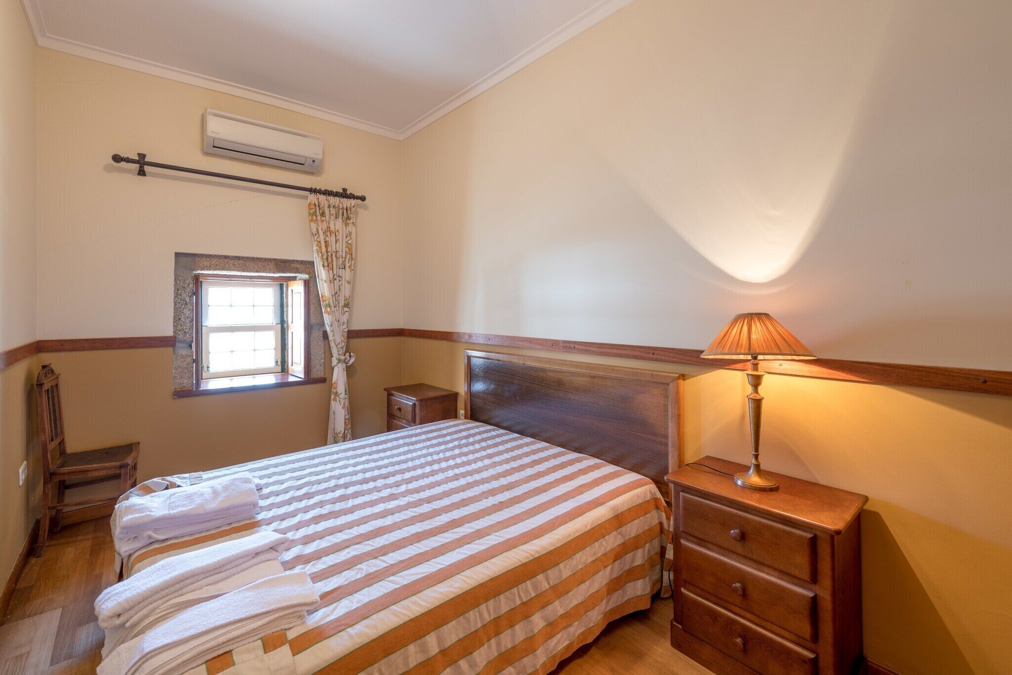 Une chambre à coucher comprenant un lit, une table de chevet, une lampe, une fenêtre avec des rideaux, une chaise en bois et un appareil de climatisation.