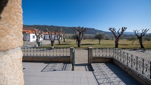 Ein gepflasterter Weg mit geometrischem Muster führt zu einem eingezäunten Bereich mit Bäumen und einer entfernten Aussicht auf Gebäude und Hügel.