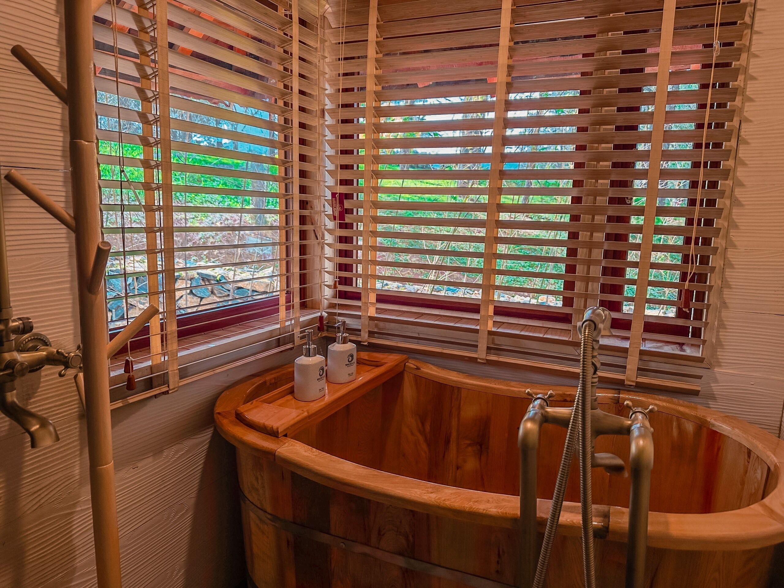 A wooden bathtub with a faucet and showerhead, wooden blinds, and a window with a view of greenery.