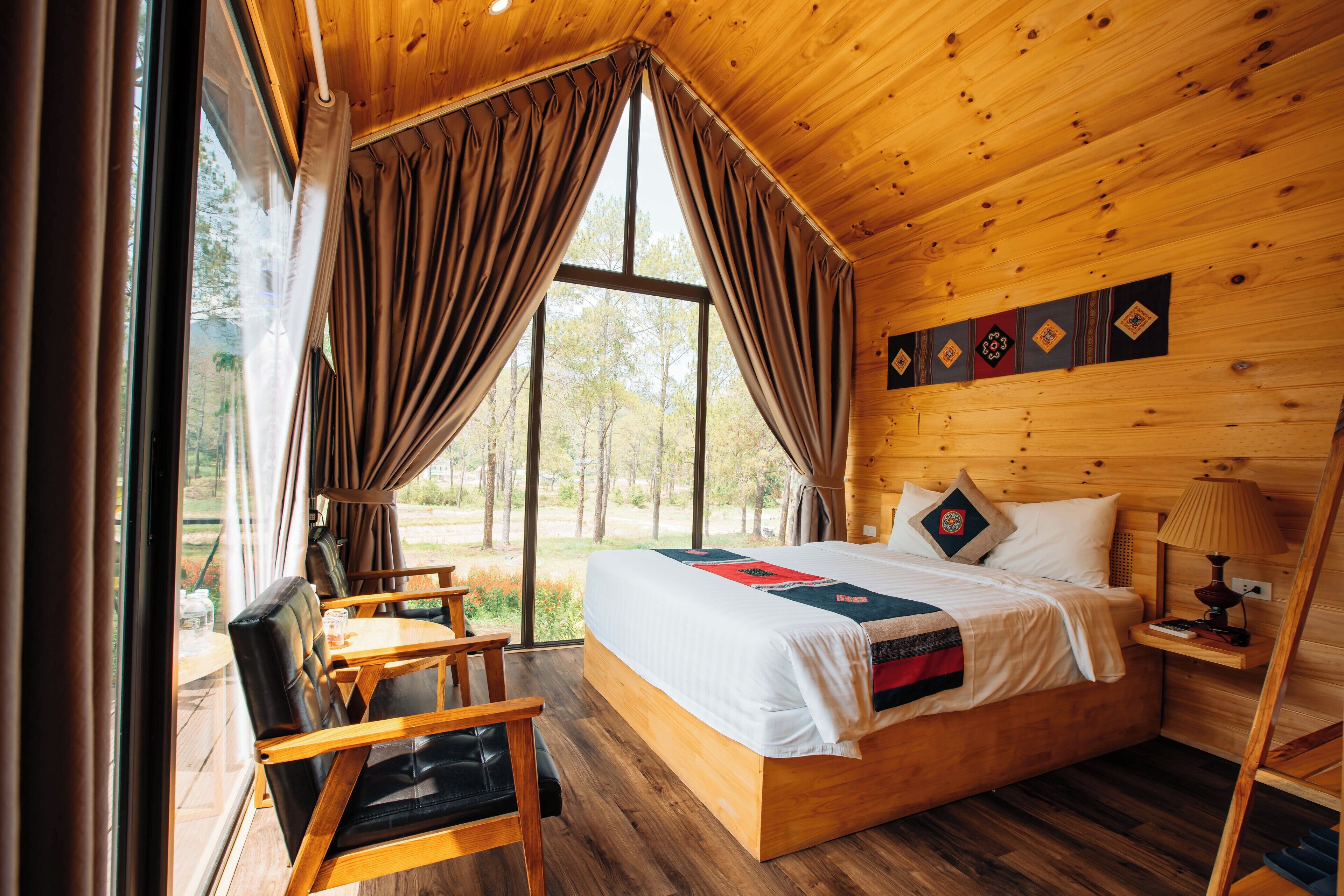 A wooden cabin bedroom with a bed, a chair, a table, and a window with curtains.