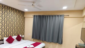 A hotel room with a bed, a ceiling fan, a window with grey curtains, a wall with a patterned wallpaper, and a TV on a wooden stand. - Emerald Tree Moshi (Pune)