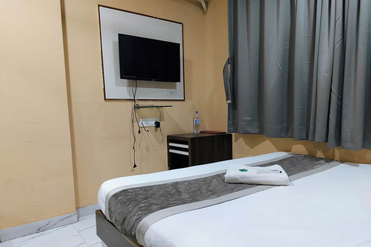 A hotel room with a bed, a flat-screen TV mounted on the wall, a small desk with a bottle of water, and a curtain on the window.
