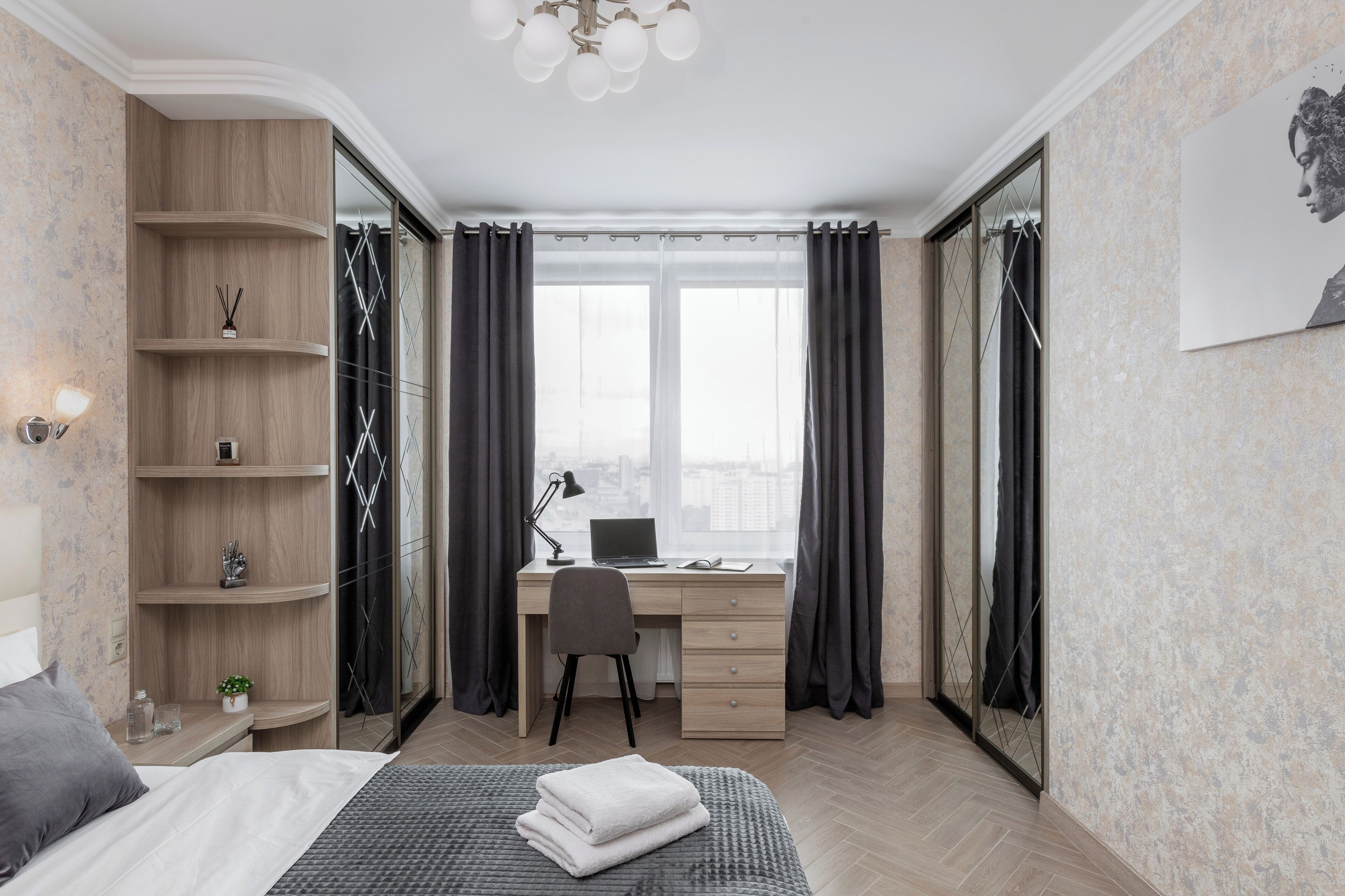A modern bedroom with a bed, a desk, and a chair. There is a window with curtains, a bookshelf, and a wall-mounted lamp.
