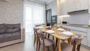 A modern dining area with a wooden table set for four, a grey sofa, and a kitchen with a tiled backsplash. - 3-room apt with view of National Library (Minsk)