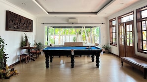 A spacious room with a pool table, a large window with a view of greenery, and a ceiling with a chandelier. - Domnak Soksan Boutique (Siem Reap)