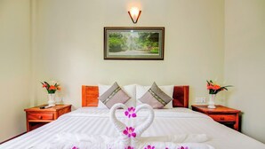 A neatly made bed with a heart-shaped towel arrangement and orchid flowers, flanked by two nightstands with vases of flowers, in a room with a framed picture on the wall. - Domnak Soksan Boutique (Siem Reap)