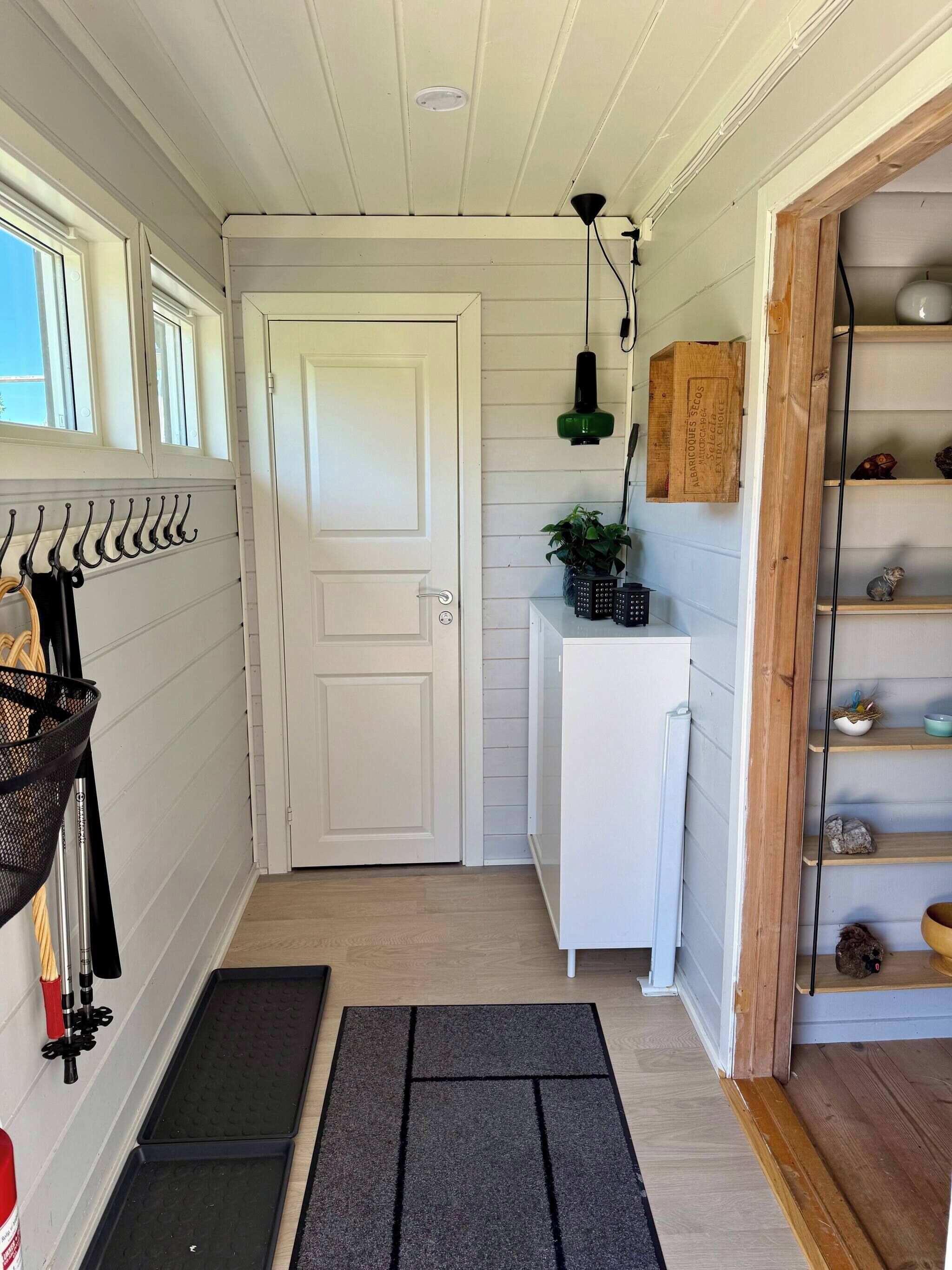 A hallway with a white door, a black mat, a shelf with decorative items, and a hanging light fixture.