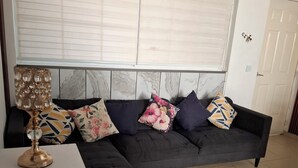 Un salón con un sofá oscuro, una mesita auxiliar blanca, una lámpara decorativa y una ventana con persianas.