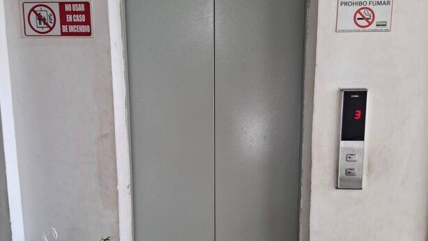 Un ascensor con una pantalla digital que muestra el número 3, un cartel encima que indica 'PISO-3', y dos señales de seguridad en la pared.