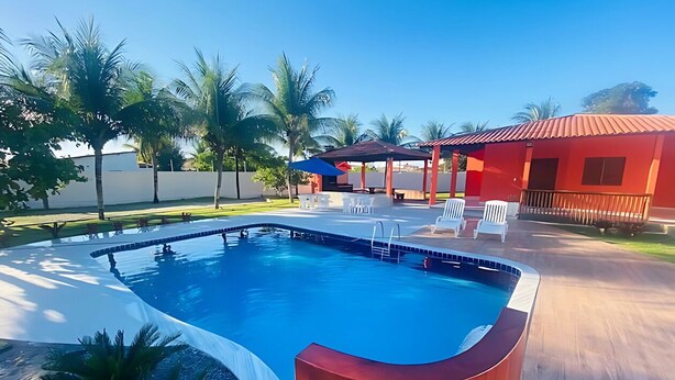 A swimming pool with a wooden deck, white lounge chairs, and a red building in the background. - BSM Beach House - Top Leisure! (Barra Mar Barra de São Miguel)