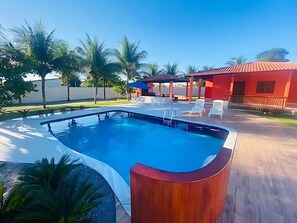 A swimming pool with a wooden deck, white lounge chairs, and a red building in the background.