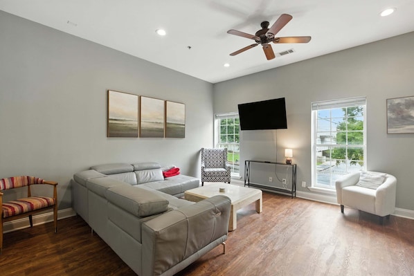 A spacious living room with a grey sectional sofa, a wooden coffee table, a patterned armchair, and a white armchair. A ceiling fan and a flat-screen TV are mounted on the wall.