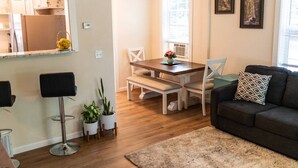A modern living room with a dark sofa, a coffee table, and a dining area with a wooden table and chairs. There are potted plants and a bar stool visible.