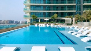 A rooftop pool with clear blue water, surrounded by white lounge chairs and umbrellas, with a modern building in the background. - Beachfront 2BR w Palm & Sea Views (Dubai)