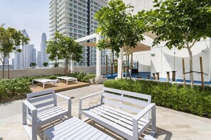 Un espace extérieur aménagé avec des bancs et des tables blancs, entouré de verdure et offrant une vue sur un paysage urbain avec de hauts immeubles.