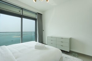 Une chambre d’hôtel avec un grand lit, une table de chevet blanche, une commode moderne et une vue sur la mer grâce à de grandes fenêtres du sol au plafond.