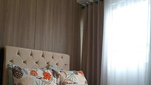 A bedroom with a tufted headboard, a bed with floral-patterned pillows, a small bedside table, and a window with sheer curtains. - COAST RESIDENCE (Pasay)