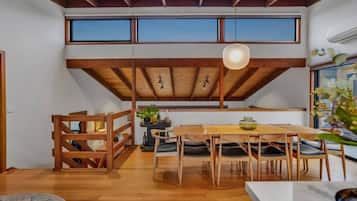 A dining area with a wooden table and chairs, a hanging light fixture, and large windows.