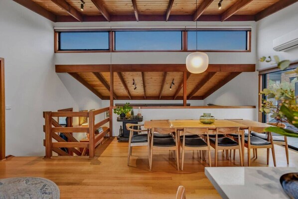 A dining area with a wooden table and chairs, a hanging light fixture, and large windows.