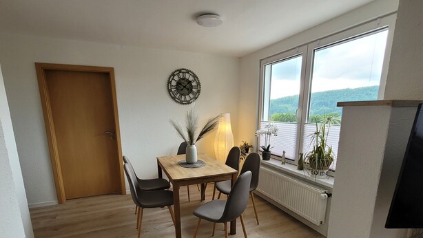 Een eetgedeelte met een houten tafel, vier stoelen, een wandklok, een lamp en een raam met uitzicht op groen.