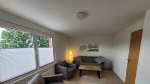 Een compacte leefruimte met een grijze bank, een kleinere fauteuil, een houten salontafel en een lamp. Een radiator is links zichtbaar, en een deur bevindt zich rechts.