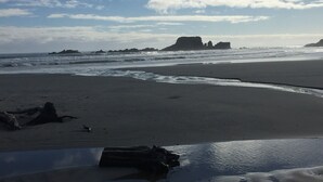 A beach with a rocky outcrop in the distance, waves approaching the shore, and a clear sky with scattered clouds.