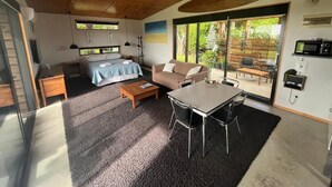 A modern living room with a sofa, coffee table, and dining table. There is a sliding glass door leading to an outdoor area with a wooden deck.