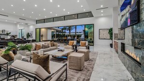 A spacious, well-lit living area with a modern design, featuring a large seating area, a fireplace, and a view of the outdoors. - Sleeps 54 | 3 Luxury Villas w/ Pool + Spa, Pickleball | Bravo by AvantStay (Indio)
