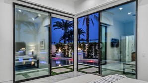 A modern room with large glass doors leading to a pool area, featuring a sofa and a dining table. - Sleeps 54 | 3 Luxury Villas w/ Pool + Spa, Pickleball | Bravo by AvantStay (Indio)