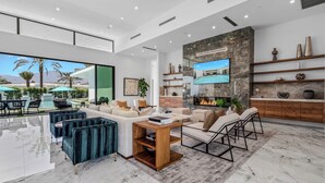 A spacious living area with a large window overlooking a pool and mountains, a fireplace, and a TV mounted on a stone wall. - Sleeps 54 | 3 Luxury Villas w/ Pool + Spa, Pickleball | Bravo by AvantStay (Indio)
