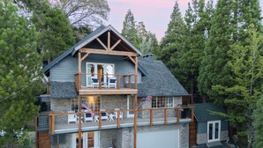 A two-story cabin with a balcony, stone exterior, and an American flag. - Sauna, Lake Views + Firepits | Near Lake Arrowhead Village | Zurich Drive (Lake Arrowhead)