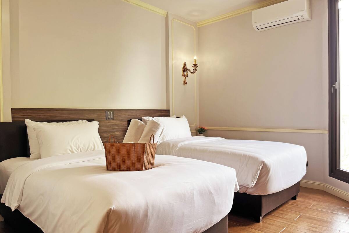 Habitación de hotel con dos camas, una cesta de mimbre sobre la cama izquierda, un equipo de aire acondicionado en la pared y una ventana a la derecha.