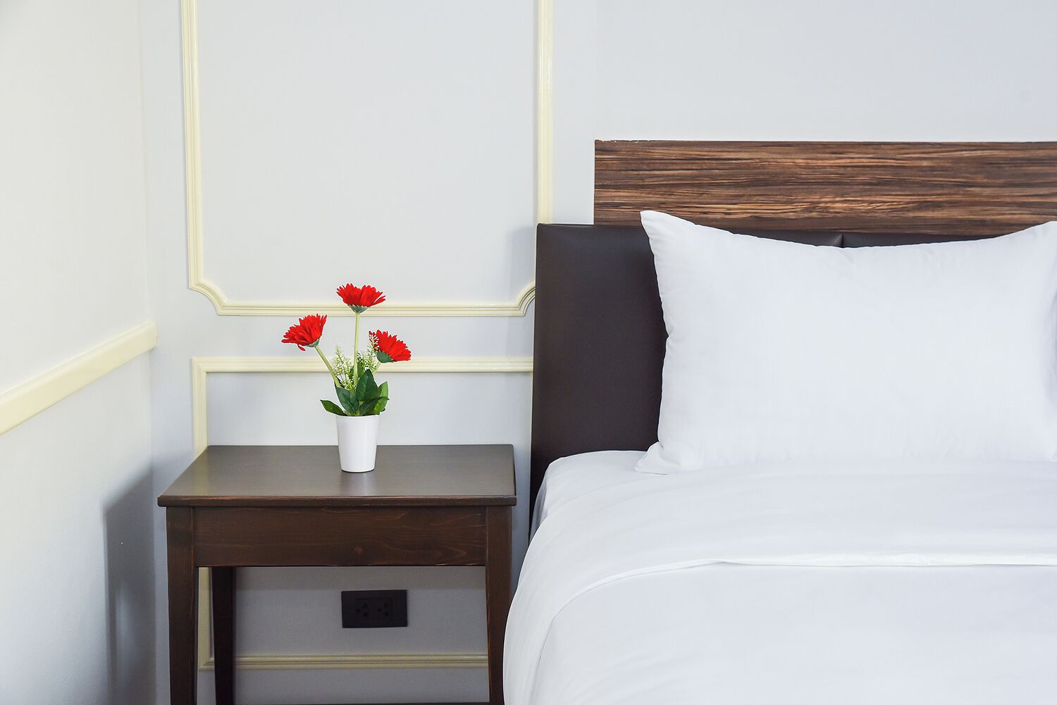 Ein ordentlich bezogenes Bett mit weißen Laken und einem dunklen Kopfteil, ein kleiner Nachttisch mit einer weißen Vase voller roter Blumen, und eine Wand mit schlichtem, klarem Design.