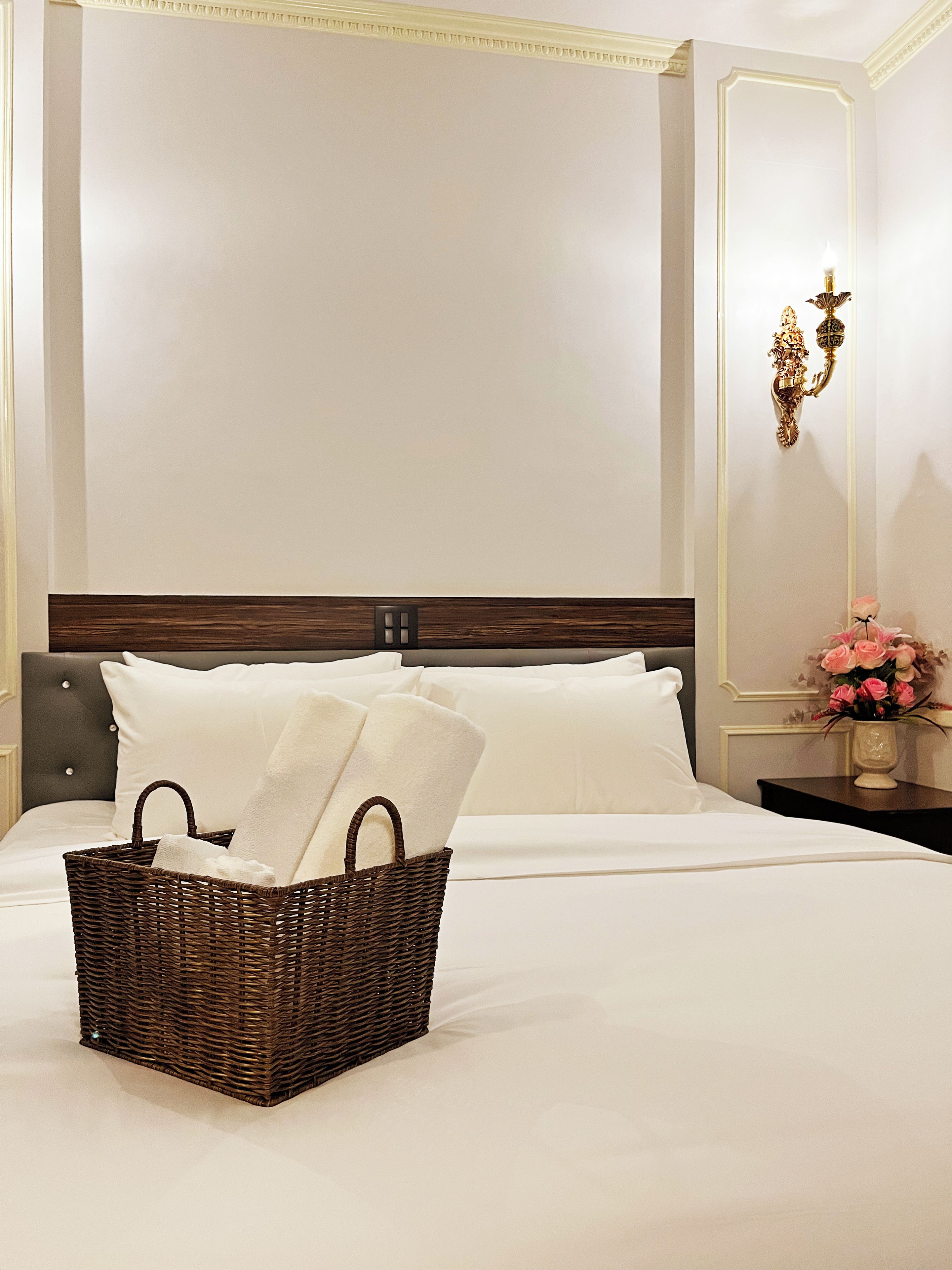 A neatly made bed with a basket of towels, a nightstand with a vase of flowers, and a wall-mounted light fixture.