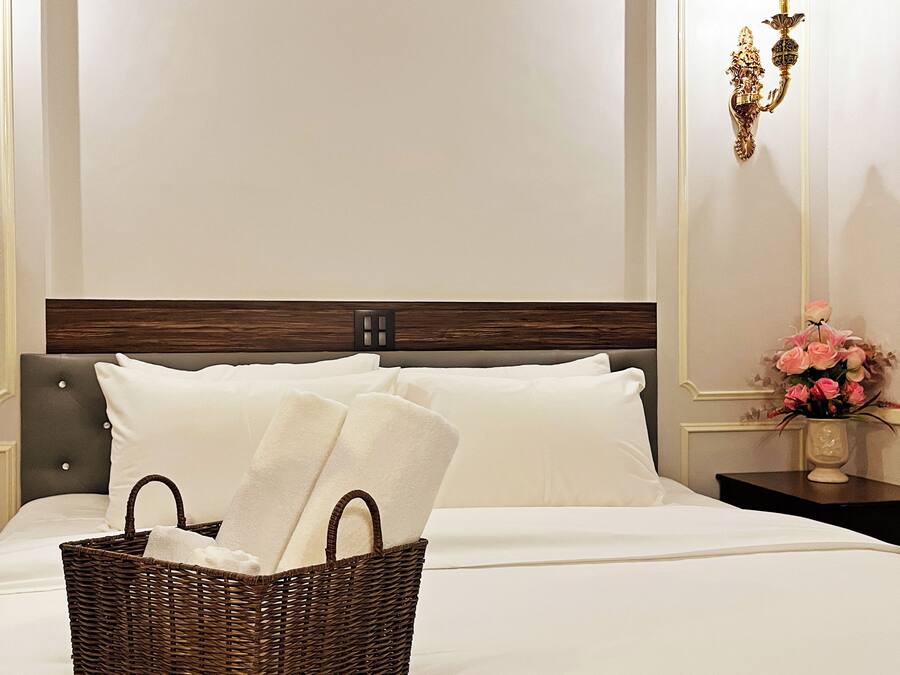 A neatly made bed with a basket of towels, a nightstand with a vase of flowers, and a wall-mounted light fixture.