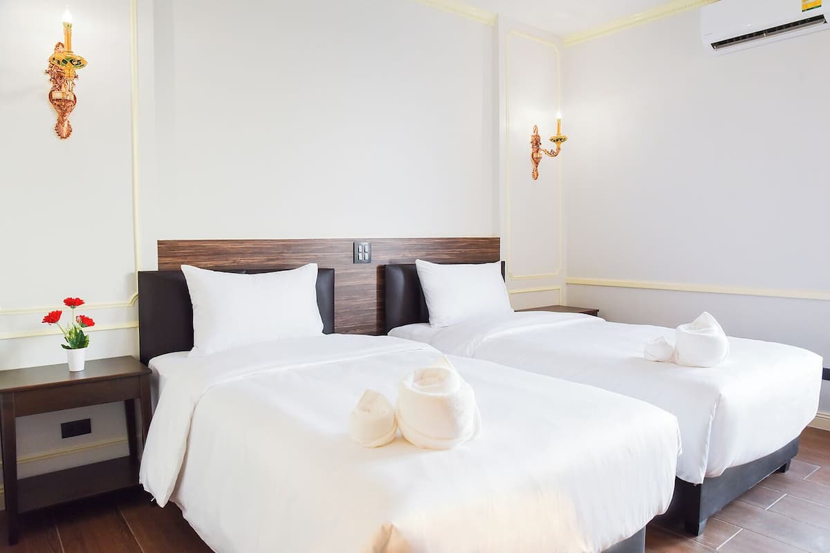 Habitación de hotel con dos camas, ropa blanca, cabecera de madera, mesita de noche con un florero de flores rojas y apliques de pared.