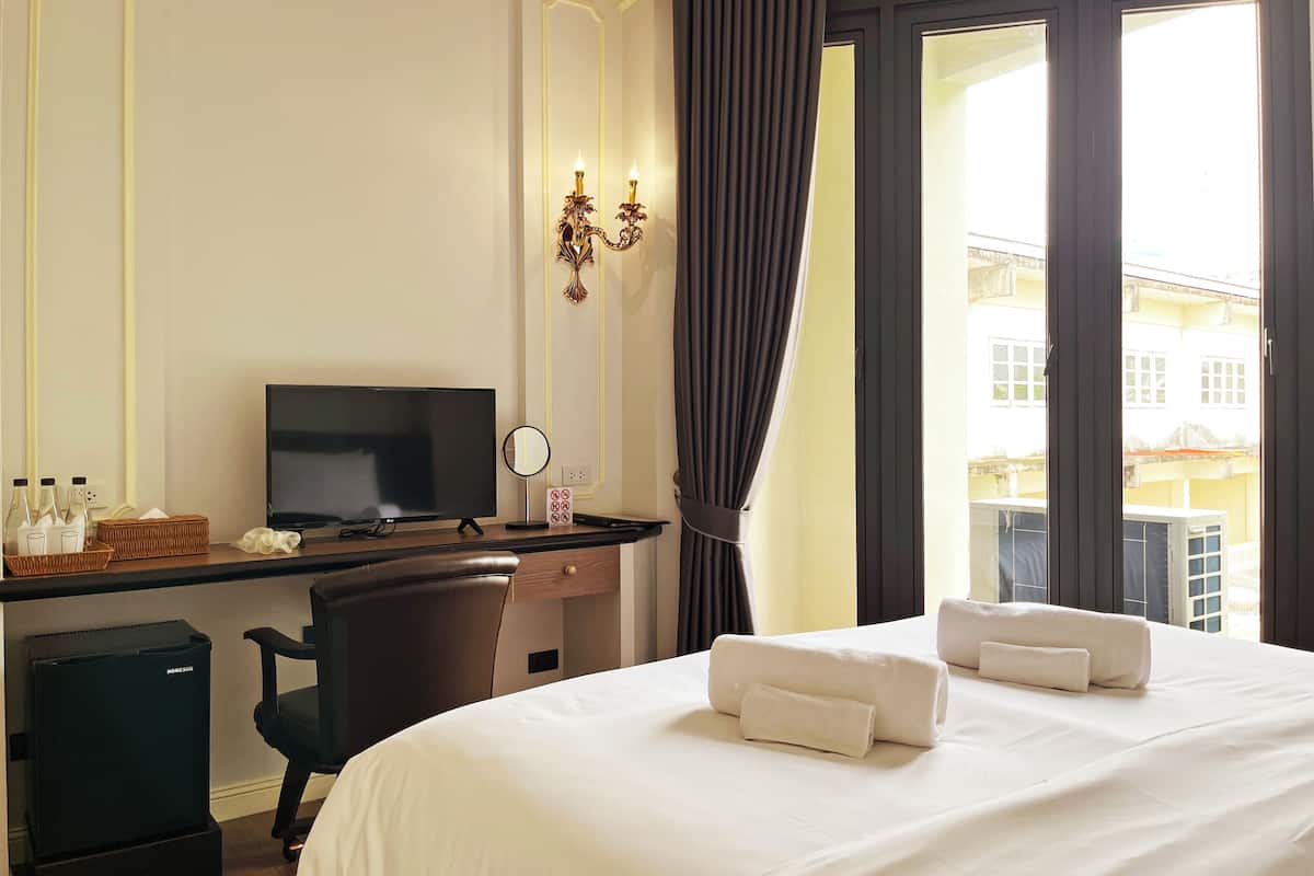 Habitación de hotel con cama, escritorio con televisor, silla, refrigerador pequeño, ventana con cortinas y vista a edificios.