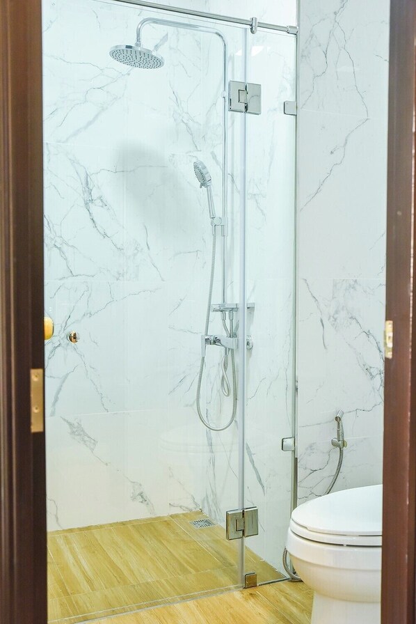 A glass-enclosed shower with a handheld showerhead and a handheld shower nozzle, a toilet, and a wooden floor.