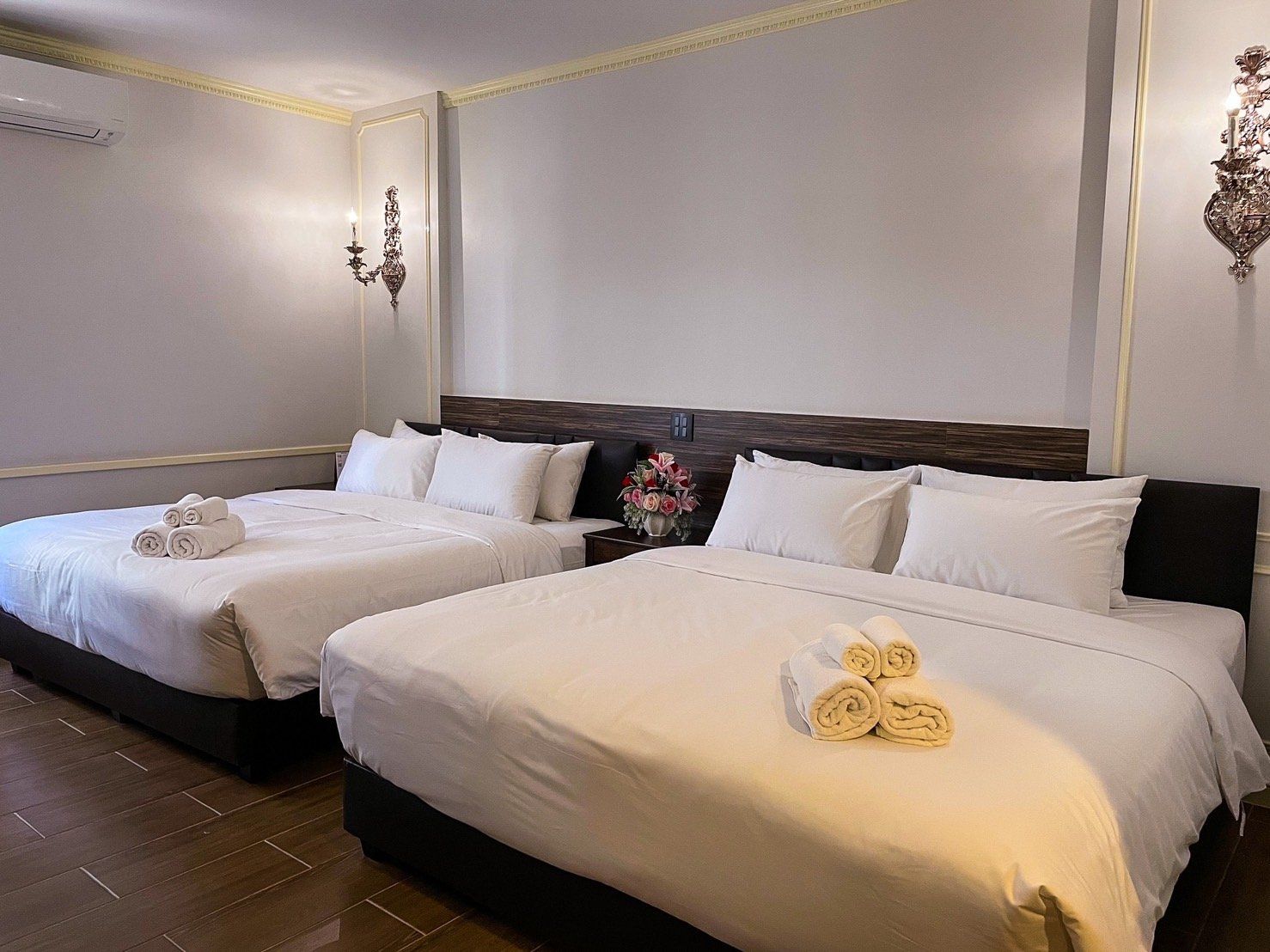 Habitación de hotel con dos camas, cada una con ropa blanca y toallas enrolladas. Hay una mesita de noche con un jarrón de flores entre las camas.