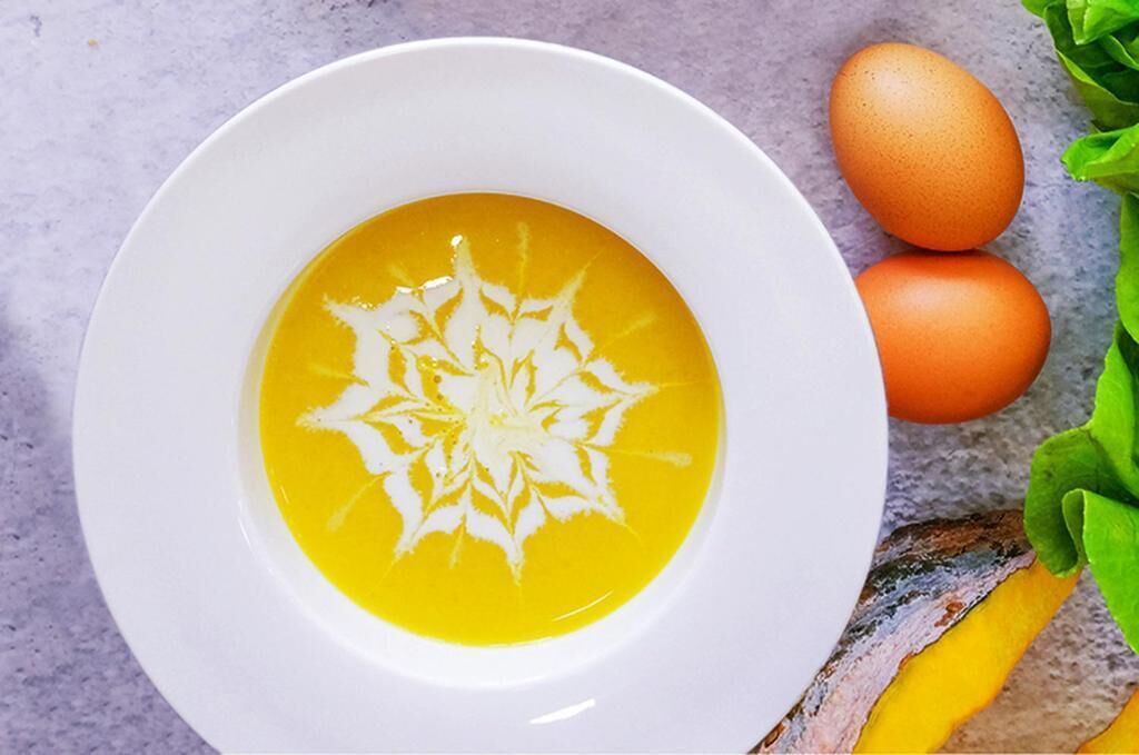 A bowl of soup with a decorative pattern on a white plate, accompanied by two eggs and some green leaves.