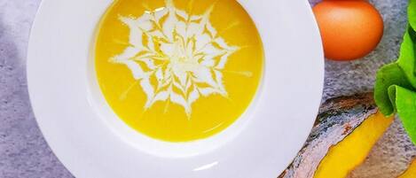 A bowl of soup with a decorative pattern on a white plate, accompanied by two eggs and some green leaves.