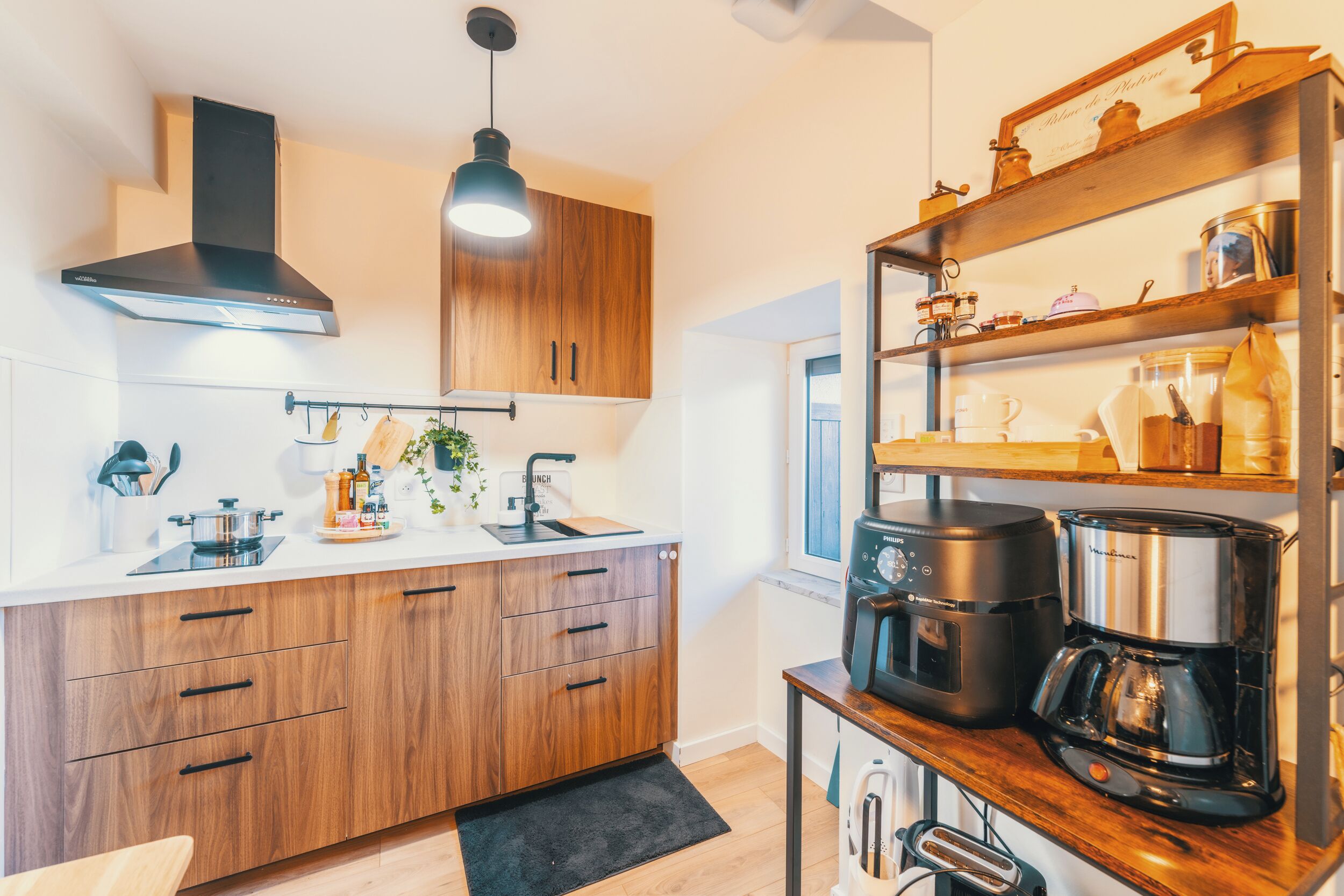 Une cuisine moderne avec des meubles de rangement en bois, une hotte noire, un évier, une machine à café et une étagère contenant des ustensiles de cuisine.