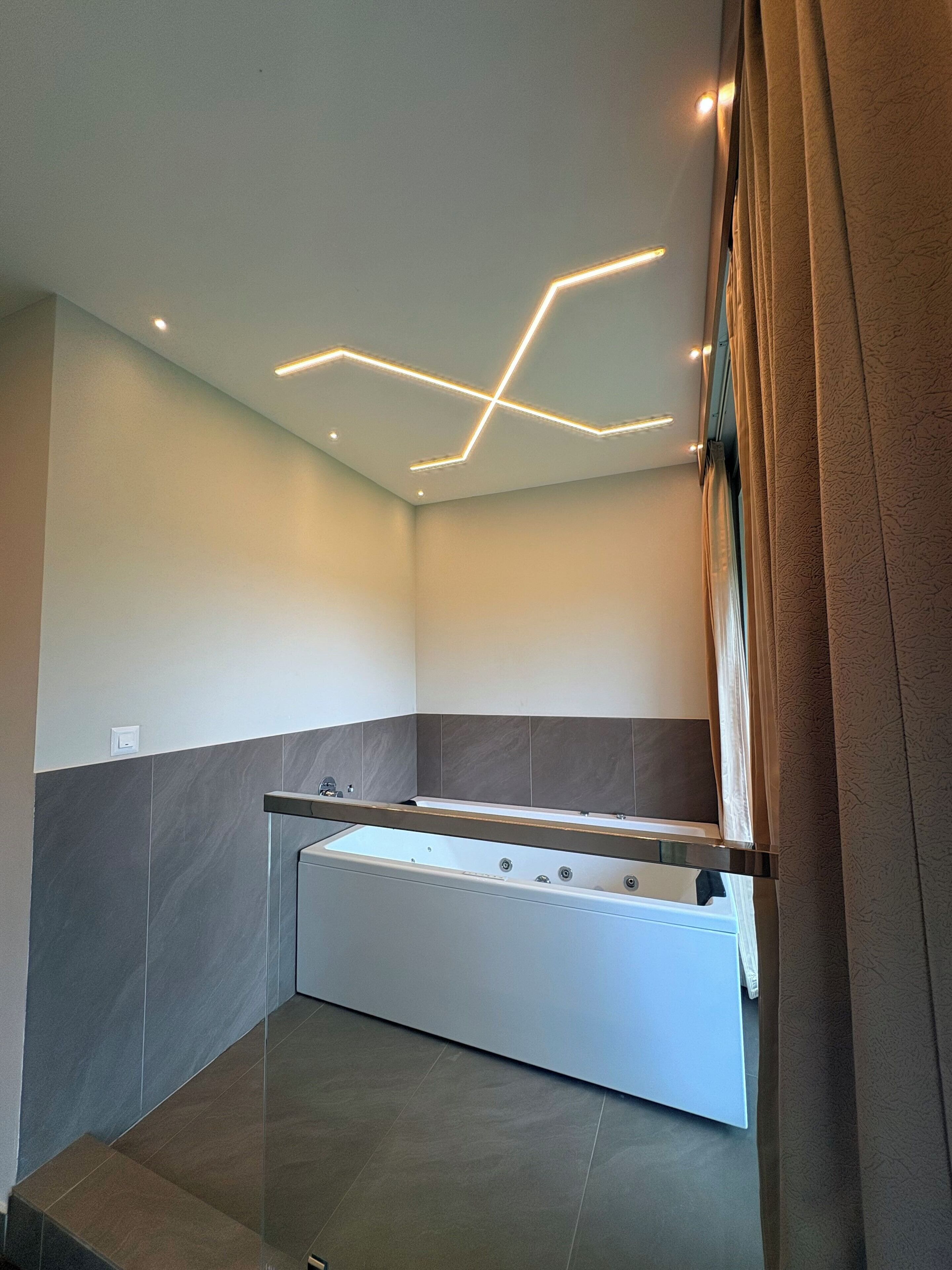 A modern bathroom with a glass-enclosed bathtub, grey and white tiled walls, and a geometric ceiling light design.
