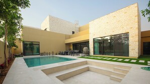 A modern outdoor pool area with a stone facade, a seating area with tables and chairs, and a clear sky. - Silk Valley - 4B  private beach access (Ghantoot)