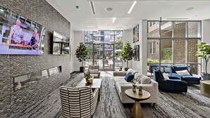 A modern hotel lobby with a large TV, comfortable seating, a patterned carpet, and large windows offering a view of the building's exterior. - CPCC Area Escape | Pool Gym Parking Balcony (Charlotte)