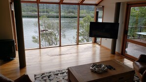 Un salon spacieux doté d’une grande fenêtre donnant sur un lac, d’un téléviseur à écran plat fixé au mur et d’une table basse en bois sur laquelle est posé un vase décoratif.